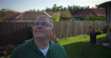 Arka bahçeye bakan bir adam ahşap çitlerin ve konut evlerinin yakınına günlük ve düşünceli bir an, banliyö mahallesi, yeşillik ve sakin bir çevre.