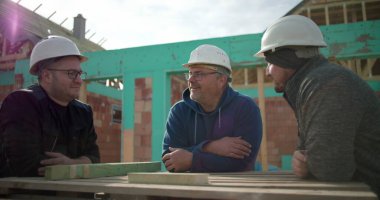 İnşaat işçileri, arka planda tamamlanmamış bir yapı iskeleti olan bir inşaat sahasında proje planlarını tartışırken tasasız bir anı paylaşıyorlar.