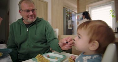 Baba kahvaltıda bebeğini yaban mersiniyle besliyor, sıcak ve sıcak bir aile ortamında aile bağlarını ve bakımı teşvik ediyor.