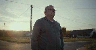 Karanlık çöktüğünde kırsal yolda duran bir adam, sakin bir ifadeyle, arka planda gün batımı parıltısı, kırsal bir manzara, huzurlu bir an, açık bir manzara, kırsal alanda yalnızlık.