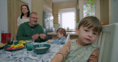 Genç bir çocuk yemek sandalyesine yaslanmış meraklı görünüyor, babası küçük çocuğu yüksek sandalyede besliyor, annesi arka planda ayakta gülümsüyor, aile mutfakta kahvaltı için toplanıyor.