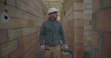 İnşaat işçisi miğfer takıyor, tamamlanmamış evin dar koridorlarında yürüyor, inşaat alanındaki tuğla duvarları ve tavan kirişlerini geçiyor.