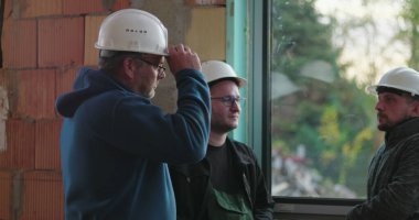 Cam kenarında sohbet eden inşaat işçileri, rahat ve günlük etkileşimler, kapalı alanda yenileme alanı, dostluk ve takım çalışması, güvenlik teçhizatı ve sert şapkalar.