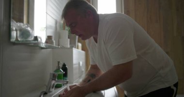 Adam lavaboda yüzünü yıkıyor, elleri suyun altında öne eğik, traş olduktan sonra dinleniyor ve dinleniyor, doğal ahşap detaylarla dolu bir banyo.