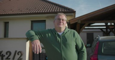 Evin önündeki iskan kapısına yaslanan bir adam, garaj yolu ve park edilmiş arabanın yanında gelişigüzel poz veriyor, banliyö ortamındaki rahat açık hava anına odaklanıyor.