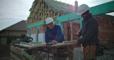 İnşaat işçisi ahşap çekiçlerken, meslektaşı inşaat alanında gözlem yapıyor, konut inşaatında takım çalışması ve el işçiliği yapıyor, arka planda ev iskeleti yapıyor.