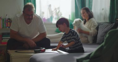 Baba sehpada çocuk ve oyuncak trenle oynuyor, anne kanepede oturuyor küçük beyaz bir köpek tutuyor, sıcak aile bağı kurma anı kapalı bir oturma odasında.