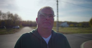 Arka planda evleri ve yeşilliği olan sakin bir banliyö sokağında güvenle gülümseyen orta yaşlı bir adam, rahat bir tavırla gelişigüzel ayakta duruyor.