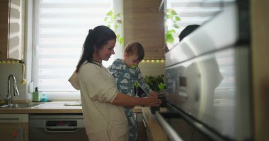Anne bebeğini mutfakta tutarken çocuk sıcak bir yuva ortamında günlük aile rutini sırasında tezgaha, bağ kurma ve ebeveynlik anına doğru uzanır..
