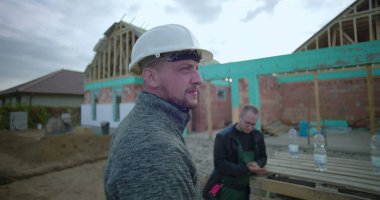 Ön planda güvenlik kaskı olan inşaat işçisi, gözlem alanı gelişimi, arka planda çalışan meslektaşı, kısmen inşa edilmiş ev yapısı görünür.