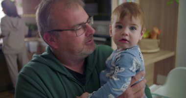 Baba mutfakta bir bebek tutuyor, çocuğa şefkatle bakıyor, düşünceli bir yüz ifadesi, sıcak ve samimi bir aile anı, arka planda yemek hazırlayan bir kadın.