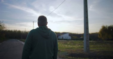 Sessiz kırsal bir yolda yürüyen, arkasını dönüp arkasına bakan düşünceli ve içe dönük, sakin bir çevre, uzaktaki evleri olan açık kırsal kesimde yaşayan bir adam.