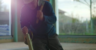 İnşaat işçisi odun parçalarını odaklama ve kuvvetle çekiçliyor, iç inşaat sahasında diz çöküyor, malzemeleri güvence altına alıyor, inşaat projesi devam ediyor.