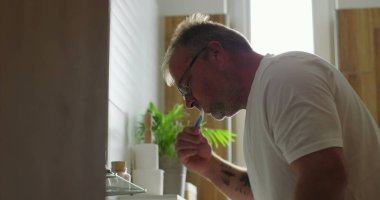 Banyo lavabosunda dişlerini fırçalayan adam, günlük sabah rutininin bir parçası olarak diş hijyenine odaklanıyor, minimalist ve temiz ortamlarda pencereden ışık yayılıyor.