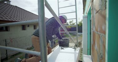 Yalıtım tahtasına yapıştırıcı süren inşaat işçisi, duvar kurulumu, güvenlik kaskı, arka planda merdiven ve yapı iskelesi olan dış yapı projesi için hazırlık yapıyor.