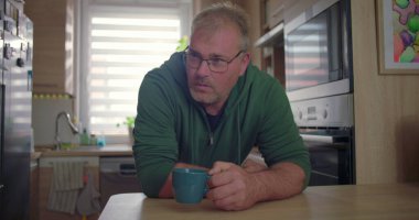 Mutfak tezgahında oturan adam, fincanda yansıtıcı pozlar, ev hayatı, modern ev ortamı, doğal ışık ve yeşillik ile günlük sabah rutini.