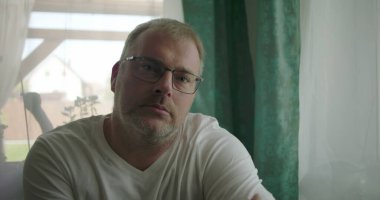 Pencerenin yanında oturan bir adam, sakin bir ev ortamında yumuşak perdelerle çevrili nötr ve içe dönük bir ifadeyle doğrudan kameraya bakıyor.