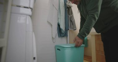 Modern bir banyodan mavi çamaşır sepetini taşıyan bir adam çamaşır makinesine bir parça çamaşır koyuyor. ev rutini