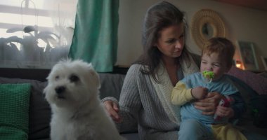 Anne kucağında bebeğe gülümserken pofuduk beyaz köpek yakınlarda oturuyor, oturma odasında neşeli bir bağ, elinde oyuncak ve emzik tutan bir bebek..