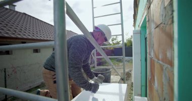 Yalıtım tahtasına yapıştırıcı süren inşaat işçisi, duvar kurulumu, güvenlik kaskı, arka planda merdiven ve yapı iskelesi olan dış yapı projesi için hazırlık yapıyor.