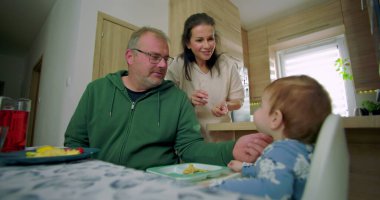 Anne ve baba yüksek sandalyede oturan bebekle iletişim kuruyor, baba bebeği besliyor, anne sıcak bir aile mutfağında gülümsüyor.