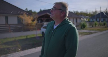 Orta yaşlı bir adam sakin bir mahallede düşünceli bir ifadeyle yukarı bakıyor, banliyö evleri ve yeşilliklerin yanında gelişigüzel duruyor.