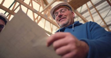 Mimari planları birlikte inceleyen inşaat işçileri, kısmen inşa edilmiş bir yapı içinde proje planlama ve tasarım planlarını analiz ediyorlar.