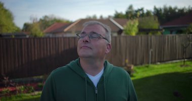 Arka bahçeye bakan bir adam ahşap çitlerin ve konut evlerinin yakınına günlük ve düşünceli bir an, banliyö mahallesi, yeşillik ve sakin bir çevre.