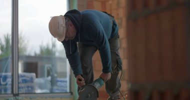 İnşaat işçisi çimento kesiyor, kıvılcımlar çıkarıyor, güvenlik kaskı yapıyor, göreve odaklanıyor, bina içi inşaata dikkat ederek ağır makineleri idare ediyor.