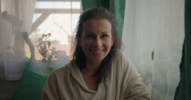 Kadın pencerenin yanında otururken sıcacık gülümsüyor, rahat ve neşeli bir yüz ifadesi sergiliyor rahat bir evin içinde, yumuşak dekorlu ve doğal ışıklandırmalı.