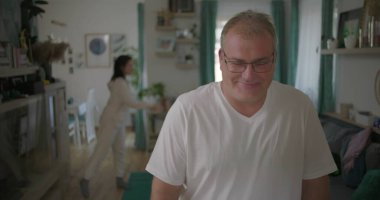 Evde hararetli tartışmalar yaşayan bir çift, erkek alaycı bir gülümseme sergilerken, kadın arka planda hayal kırıklığı içinde, evde gerginlik ve duygusal kopukluk sergiliyor.