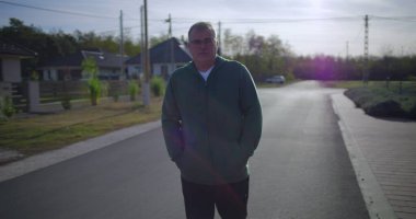 Orta yaşlı bir adam sakin bir mahallede düşünceli bir ifadeyle yukarı bakıyor, banliyö evleri ve yeşilliklerin yanında gelişigüzel duruyor.