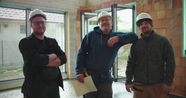 Tamamlanmamış binada inşaat ekibi, proje planlarını ellerindeki planlar, takım çalışması, güvenlik kaskları, işbirliği ve inşaat ayrıntılarına odaklanıyor.