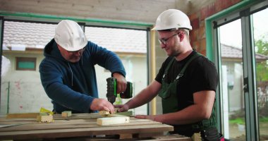 İnşaat işçileri inşaat sahasında ahşap döşeme, yapı iskelesinde takım çalışması, bir işçi sondajı, diğeri asistanlar, hassasiyet ve işbirliğine odaklanıyor.