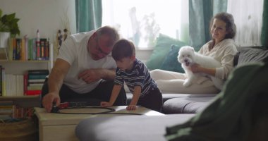 Aile, sıcak oturma odasında birlikte vakit geçirir. Büyükbaba, küçük çocukla oynar. Anne, evcil köpekle kanepede gülümser.