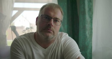 Pencerenin yanında oturan bir adam, sakin bir ev ortamında yumuşak perdelerle çevrili nötr ve içe dönük bir ifadeyle doğrudan kameraya bakıyor.