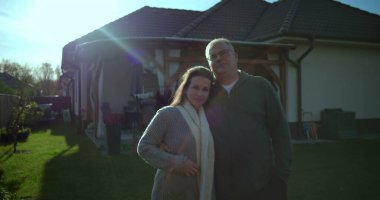 Arka bahçelerinde kucaklaşan bir çift, gururlu ev sahipleri olarak samimiyet ve iletişim içinde, arka planda bakımlı banliyö evleri görünür.