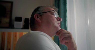 Pencere kenarında duran bir adam, çeneye yakın, derin düşüncelere dalmış, gözlük takmış, yansıtıcı ifade takmış, hayatı kapalı bir yerde düşünüyor, sakin bir an, içe kapanık ve sakin bir ruh hali.