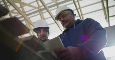 İnşaat denetçisi inşaat planlarını gözden geçirirken aynı zamanda inşaat çalışanı, takım çalışması ve yapısal proje rehberliği, güvenlik kaskları takılı
