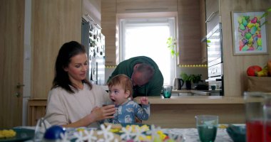 Anne bebeğe içkiyle yardımcı olurken baba mutfak tezgahına yaslanıyor, büyük çocuk sıcak aile ortamında sıcak bir yemek masasında babasıyla etkileşime geçiyor.