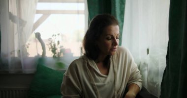 Kanepede oturan bir kadın, elleri kucağına yapışık, penceresinden dışarı bakan düşünceli bir yüz ifadesiyle, rahat bir ev içi ve yumuşak bir dekorla çevrili.