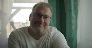 İçerde sıcak bir şekilde gülümseyen bir adam, kolları çaprazlanmış bir pencerenin yanında oturan, parlak ve sakin bir ev ortamında dostluk ve sakinlik saçan bir adam.