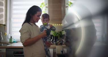 Anne mutfakta gülümserken bebeği kucağına alıyor, günlük ebeveynlik rutini sırasında çocukla meşgul oluyor, ev ortamında sıcak ve sıradan bir aile sahnesi..