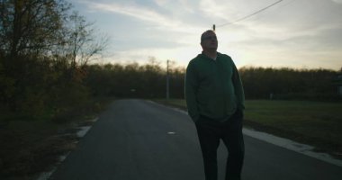 Gün batımında boş bir kırsal yolda yürüyen bir adam, elleri ceplerinde, huzurlu bir kırsal bölgede sonbahar ağaçları ve açık gökyüzü ile huzurlu bir akşamın tadını çıkarıyor.