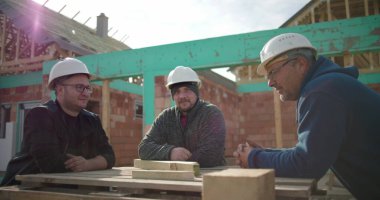 İnşaat işçileri çalışma masasının etrafında birlikte gülüyorlar, başlık takıyorlar, neşeli bir anı paylaşıyorlar, ev inşaatı sırasında bağ kuruyorlar.
