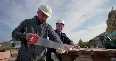 İnşaat işçisi ahşap tahtayı el testeresiyle kesiyor, iş arkadaşı gözlemliyor, inşaat sahasında takım çalışması, ev iskeleti görünür, hassasiyet ve işçiliğe odaklanmış.