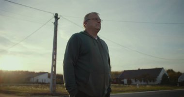 Karanlık çöktüğünde kırsal yolda duran bir adam, sakin bir ifadeyle, arka planda gün batımı parıltısı, kırsal bir manzara, huzurlu bir an, açık bir manzara, kırsal alanda yalnızlık.