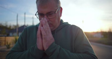 Elleri duada birbirine kenetlenmiş bir adam, kapalı gözler, derin düşüncelere dalmış ve ruhani bir an, kırsal bir arka plan, sakin ve sakin, günbatımı atmosferi