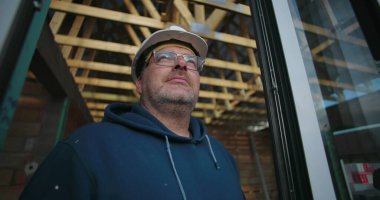 İnşaat işçisi açık çatı çerçevesine bakıyor, başlık ve gözlük takıyor, düşünceli bir ifadeyle bina inşaatının ilerlemesine odaklanıyor.