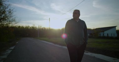 Orta yaşlı bir adam gün batımında kırsal bir yolda yürüyor, elleri ceplerinde, yansıtıcı ifade, huzurlu kırsal manzara, elektrik hatları, sakin alacakaranlık, kırsal ortam